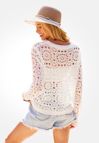 Suéter de ganchillo blanco con patrones florales y tramas abiertas, combinado con pantalones cortos de mezclilla. Llevado con un sombrero de paja con una banda.