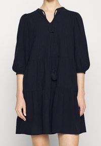 Robe noire en lin avec un col en V, des manches ballons et une jupe à volants. Elle est dotée d'une fermeture à lacets avec des pompons et de coutures verticales discrètes.