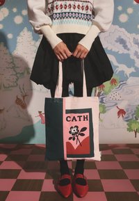 Drobės nešiojimo maišelis rožinės, žalios ir kreminės spalvų su gėlių raštu ir tekstu „CATH“. Modelis dėvi juodą sijoną, baltą megztinį ir raudonus batelius.