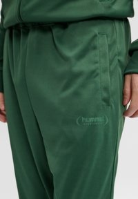 Grønne sportsbukser i glat stof, med en sidelomme og et diskret "hummel SPORTSWEAR" logo på låret.
