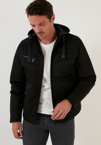 Svart zip-up jacka med huva, med texturerad panel, två fickor på bröstet med dragkedja och en slät kropp. Bärs över en vit skjorta.