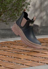 Μαύρο λαστιχένιο μποτάκι αστραγάλου με ροζ λεπτομέρειες, στρογγυλή μύτη και χοντρή σόλα· λογότυπο μπροστά και ταινία τράβηγματος πίσω.