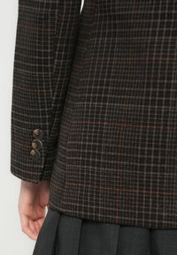Mouw van een donker geruite blazer met drie knopen, gedragen over een hand en gecombineerd met geplooide donkergrijze broek.
