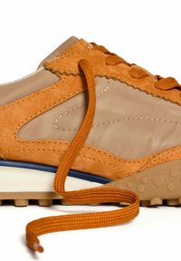 Sneaker in suede e tessuto color beige con lacci arancioni, suola testurizzata e accento blu a contrasto sulla suola intermedia. Il laccio è parzialmente slacciato.