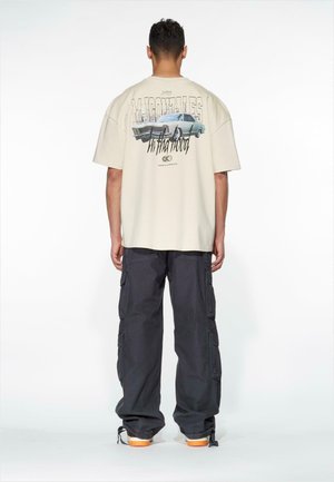 Beige oversized t-skjorte med et grafisk trykk av en bil på ryggen, kombinert med mørke cargo-bukser og sneakers med oransje såler.