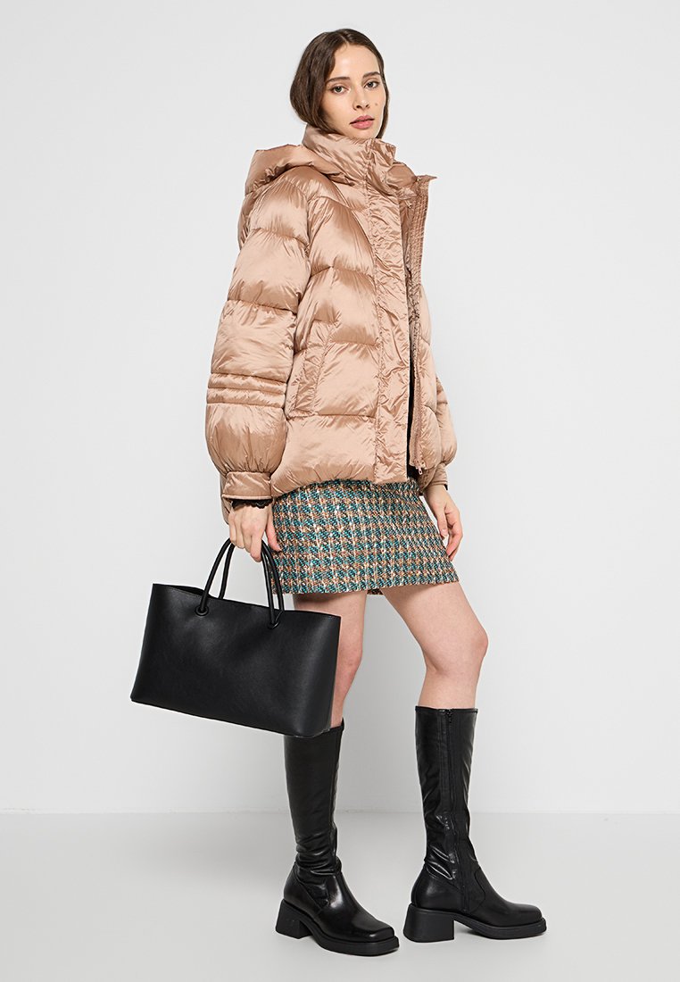Kurtka puchowa w jasnym beżu, z wysokim kołnierzem i pikowanym wzorem. Modelka nosi wzorzystą spódnicę i ma na ramieniu czarną torbę typu tote.