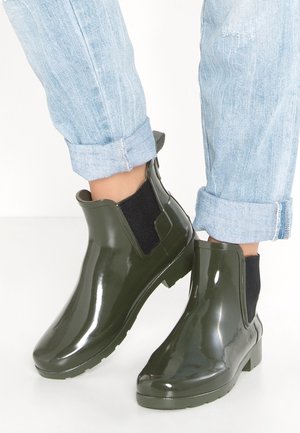 Bottes de pluie chevilles vert foncé brillant avec panneaux latéraux élastiques noirs portées avec jean bleu clair retroussé sur fond blanc.