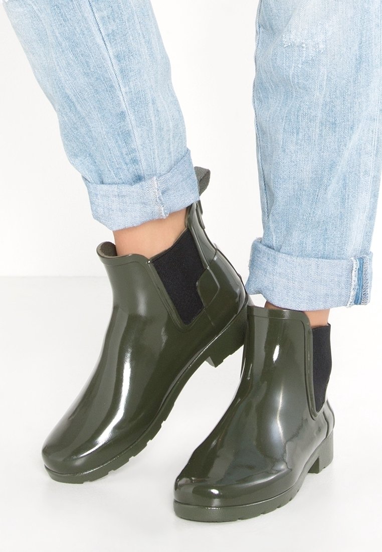 Bottes de pluie chevilles vert foncé brillant avec panneaux latéraux élastiques noirs portées avec jean bleu clair retroussé sur fond blanc.