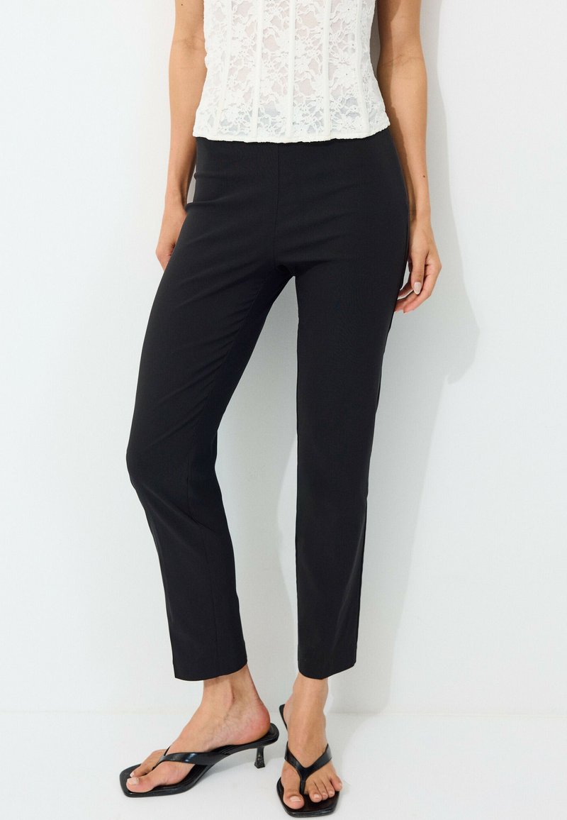 Femme portant un pantalon noir ajusté aux chevilles, un haut blanc en dentelle sans manches, et des sandales noires à talons, debout sur fond blanc.