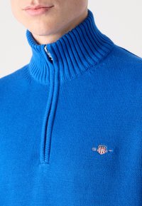 Blauer Strickpullover mit einem gerippten hohen Kragen und halbem Reißverschluss. Mit einem kleinen gestickten Logo auf der Brust. Weicher Stoff.