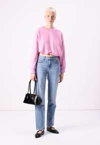 Sweatshirt cortado rosa, jeans de cintura alta azul claro, mala preta e sapatos rasos pretos com detalhes metálicos, sobre um fundo liso.