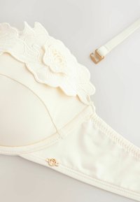 Bh i krämfärg med en broderad blommönster vid övre kanten. Har en slät tyg, justerbar rem och detaljer i guld.
