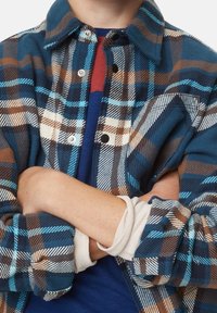 Person med korslagte arme iført en blå, brun og hvid ternet jakke over en marineblå og rød skjorte.