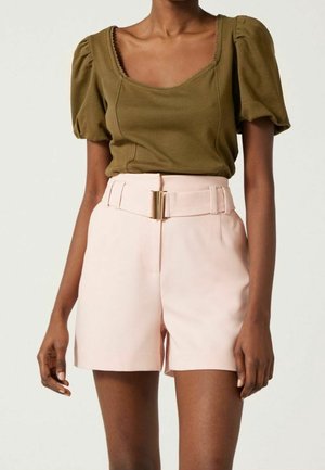 Kobieta w oliwkowej bluzce z bufiastymi rękawami oraz jasnoróżowych szortach z wysokim stanem, szerokim paskiem i złotą klamrą, pozująca na jednolitym tle.