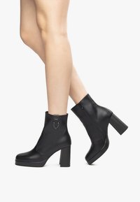 Schwarze Knöchelschuhstiefel mit einem klobigen Blockabsatz, elegantem Kunstledermaterial, seitlichem Reißverschluss und dekorativem Kettendetail am Knöchel.