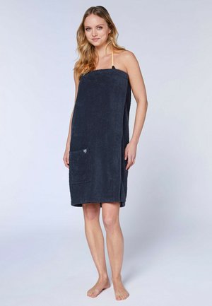 Frau trägt ein dunkeblaues trägerloses Handtuchkleid mit einer Fronttasche, steht barfuß vor einem schlichten hellen Hintergrund.