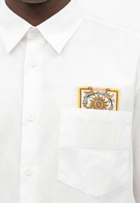 Chemise blanche boutonnée avec un écusson brodé sur le thème du soleil partiellement visible dans la poche poitrine gauche.