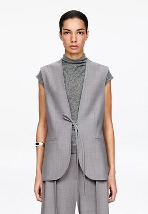 Femme portant un blazer sans manches gris noué à la taille sur un col roulé gris à manches courtes, avec un pantalon large assorti et un bracelet argenté.