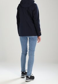 Námořnická modrá nepromokavá bunda s kapucí, s hladkou texturou a logem. Nosená přes světle modré slim džíny a černé tenisky.