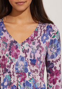 Blommig klänning med V-ringning, långa ärmar och samlade texturerade paneler i nyanser av lila, blå och grön.