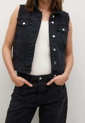 Persona que lleva un chaleco vaquero negro sin mangas con botones plateados sobre una camiseta blanca sin mangas, combinado con vaqueros negros de cintura alta.