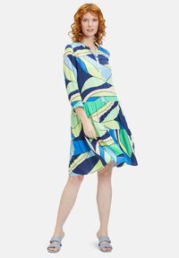 Kleid mit kurzen Ärmeln, das ein blaues und grünes Blättermuster zeigt, mit einer geknöpften Ausschnitt und einem gestuften Rock für zusätzlichen Volumen. Der Stoff wirkt glatt.
