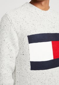 Weißer Strickpullover mit schwarzen Sprenkeln. Besitzt ein auffälliges rechteckiges Tommy Hilfiger-Logo in Marineblau, Weiß und Rot auf der Brust. Rundhalsausschnitt.