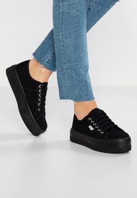 Baskets compensées en suède noir avec une semelle texturée, un design à lacets et des œillets en argent. Portées avec un jean en denim bleu effilé.