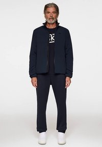 Laivastonsininen vetoketjullinen takki mustan t-paidan päällä, yhdistetty samanvärisiin laivastonsinisiin housuihin ja valkoisiin tennareihin. Sileä pinta, rento malli, vähäiset yksityiskohdat.