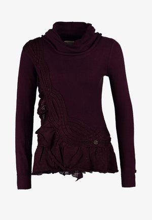 Kvinde iført en mørklilla sweater med rullekrave, stramme blå jeans, sorte ankelstøvler, der holder en sort tote-taske, stående foran en ensfarvet baggrund.