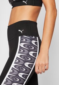 Zbliżenie na osobę w czarnych legginsach Puma z wysokim stanem i wzorzystymi panelami bocznymi w odcieniach szarości, bieli i czerni.