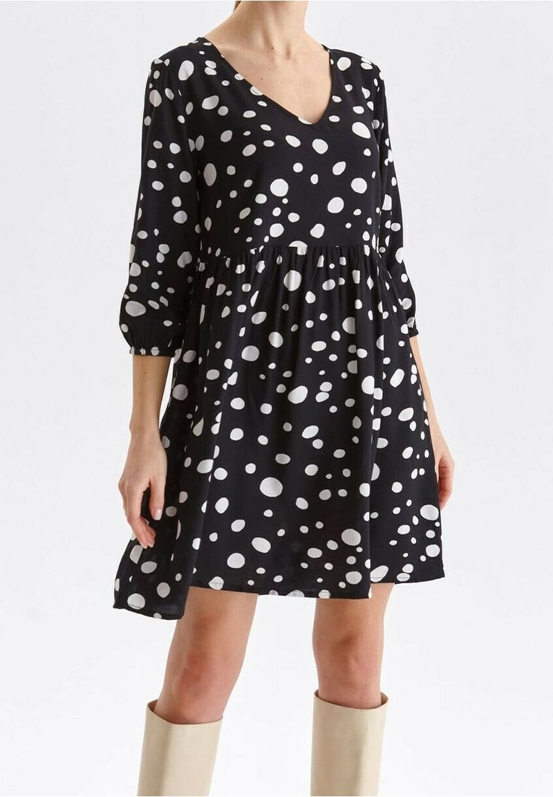 Femme portant une robe noire midi avec des pois blancs irréguliers et des manches 3/4, associée à des bottes beiges jusqu'au genou.