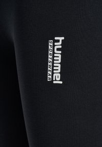 Leggings de sport noirs avec un logo "hummel" blanc et le texte "SPORTSWEAR" sur le côté gauche. Texture lisse et design ajusté.