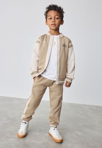 Veste en polaire beige avec boutons et détails blancs, assortie à un pantalon en polaire de la même couleur. Baskets crème avec semelles marron, dotées de lacets.