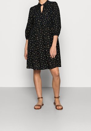 Kobieta w czarnej, kwiecistej sukience do kolan z rękawami do łokcia i brązowych płaskich sandałach z paskiem wokół kostki, stojąca na tle jednolitego tła.