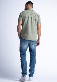 Korte mouwen olijfgroene shirt met een gebogen zoom, gecombineerd met lichtblauwe spijkerbroek en witte sneakers, gezien vanuit de achterkant.