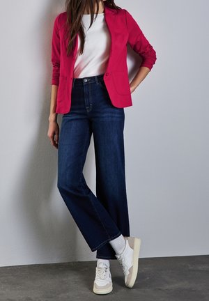Blazer rouge au design ajusté sur une chemise blanche, associé à un jean en denim foncé à jambes larges et des baskets blanches. Texture lisse.