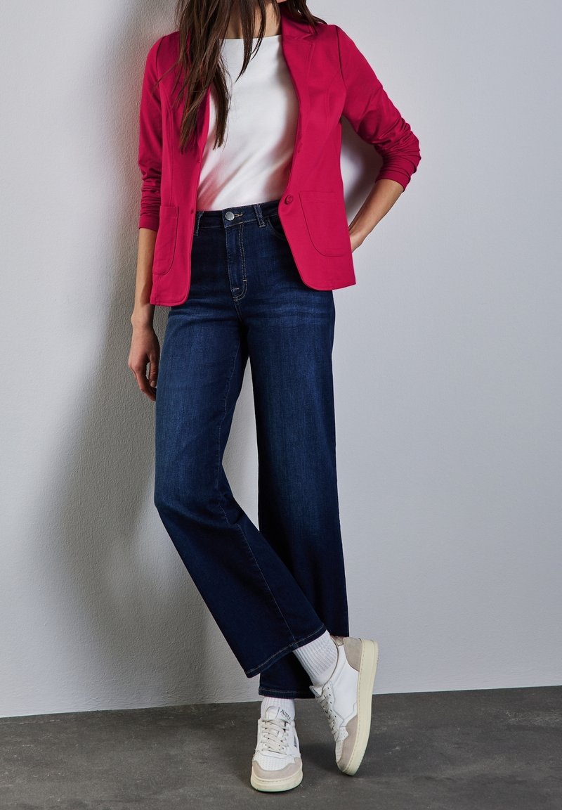 Červený blazer s přiléhavým střihem na bílém tričku, doplněný širokými tmavými džíny a bílými teniskami. Hladká textura.
