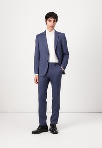 Traje azul a medida con blazer de dos botones, pantalones de corte plano y camisa blanca. Tejido suave, corte ajustado, combinado con zapatos negros.