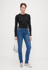 Fekete, hosszú ujjú body, kék, slim-fit farmernadrág, ezüst lapos cipők és egy fényes ezüst kézitáska. A modell egy sima fehér háttér előtt áll.