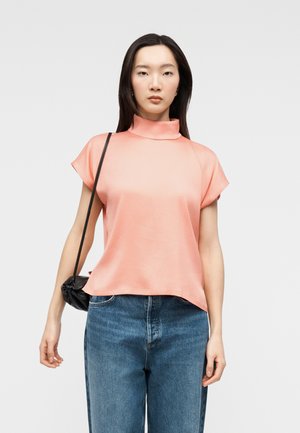 Žena v lososově růžové krátké haleně s vysokým límcem, volných modrých džínách a s malou černou kabelkou přes rameno.