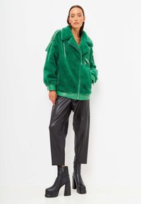 Giacca in pelliccia sintetica verde con chiusura a zip, ampio colletto e tasche con zip, abbinata a pantaloni in pelle sintetica nera e stivali neri con tacco.