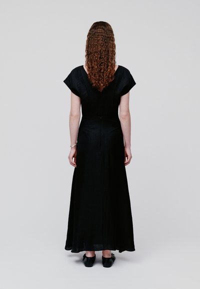 Γυναίκα με μακριά σγουρά μαλλιά, ντυμένη με μαύρο φόρεμα μέχρι τους αστραγάλους και μαύρα παπούτσια, όρθια με την πλάτη να κοιτά προς ένα απλό φόντο.