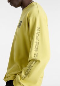 Yellow long-sleeve shirt made of soft fabric. Features a logo on the front and text "MADE FOR THE ELEMENTS" on the left sleeve.