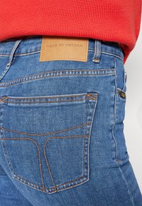 Džínové kalhoty ve světle modré barvě s oranžovým prošíváním, koženou nášivkou označenou "TIGER OF SWEDEN" a přiléhavým střihem.