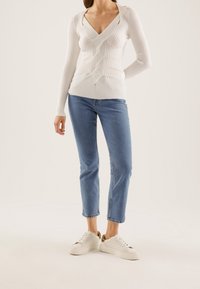 Biała sweter o żebrowanym wykończeniu, z długim rękawem i dekoltem w kształcie V, połączony z niebieskimi dżinsami o prostych nogawkach oraz białymi trampkami z złotymi akcentami.