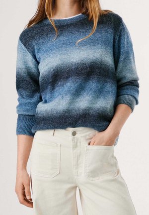 Frau trägt einen blau verlaufenden Pullover mit hochgekrempelten Ärmeln und cremefarbenen hochtaillierten Hosen, eine Hand in der Tasche, vor einem schlichten Hintergrund.