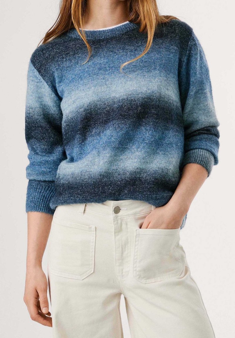 Femme portant un pull bleu dégradé à manches retroussées et un pantalon taille haute écru, une main dans la poche, sur un fond uni.