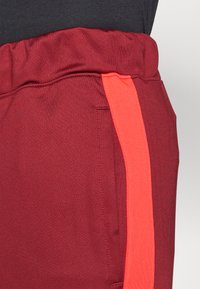 Pantalons joggers bordeaux avec une texture douce, dotés d'une large taille élastique et d'une bande latérale orange contraste.