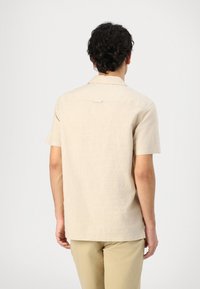 Beige kortermet skjorte laget av teksturert stoff med klassisk krage og en enkelt bakfold, brukt med lyse bukser.